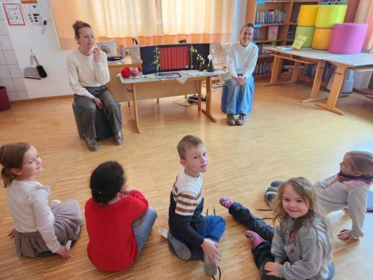 Mehrere Kinder sitzen im Kreis auf dem Boden eines hellen Raums, zwei Erwachsene sitzen an einem Tisch mit Weihnachtsdekoration.