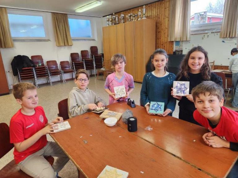 Sechs Kinder sitzen an einem Holztisch in einem Raum mit Holzvertäfelung und zeigen selbstgebastelte Sterne aus Holz und Fäden.