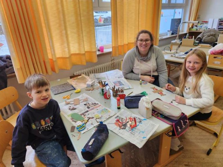 Drei Personen sitzen an einem Tisch mit Bastelmaterialien in einem hellen Raum mit gelben Vorhängen