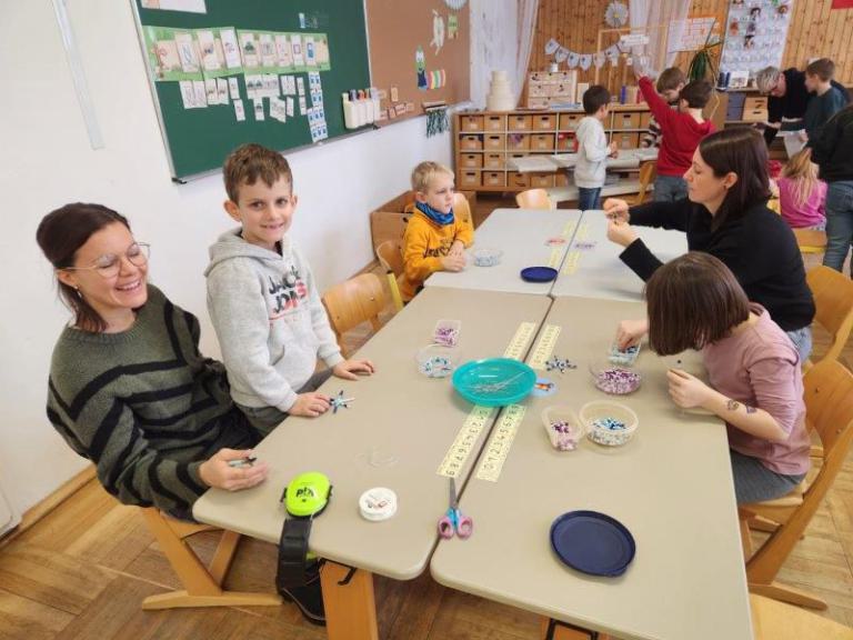 Kinder sitzen an einem langen Tisch und fädeln bunte Perlen auf Schnüre auf