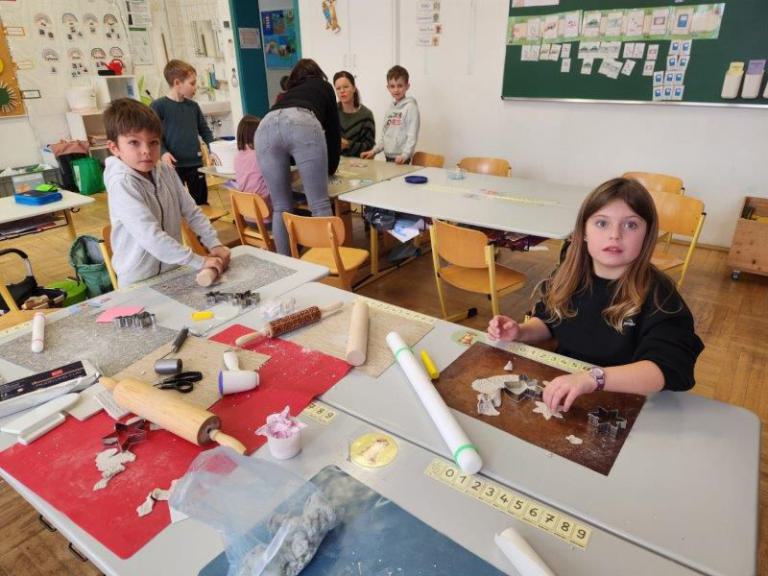 Kinder sitzen an Tischen mit Backutensilien und ausgestochenen Teigfiguren in einem Klassenraum mit Lernplakaten an der Wand