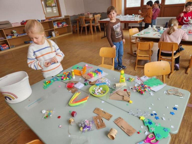Kinder basteln an einem Tisch mit bunten Bastelmaterialien wie Sternen, Pompons, Pfeifenreinigern und Kleber in einem Klassenraum.