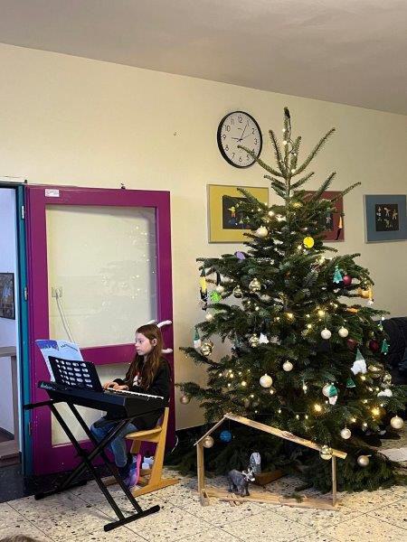 Weihnachtsbaum mit Kugel- und Lichterketten neben einer Krippe, links sitzt ein Kind an einem Keyboard.