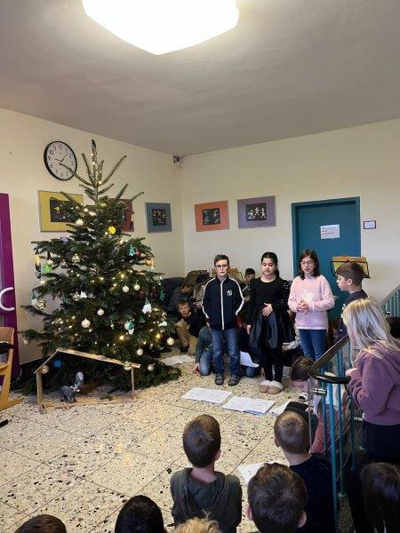 Ein Weihnachtsbaum mit Kugeln und Lichterkette davor stehen mehrere Kinder, einige halten Zettel in der Hand.