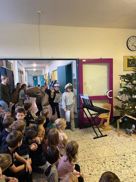 Gruppe von Kindern sitzt und steht in einem Flur, einige blicken auf einen geschmückten Weihnachtsbaum und ein Keyboard auf einem Ständer.