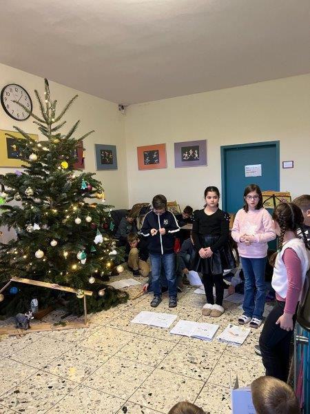 Gruppe von Kindern steht vor einem geschmückten Weihnachtsbaum in einem Raum mit bunten Bildern an den Wänden.