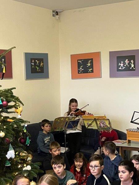 Mehrere Kinder sitzen vor einem Weihnachtsbaum, eine Person spielt Geige, bunte Kinderzeichnungen hängen an der Wand.