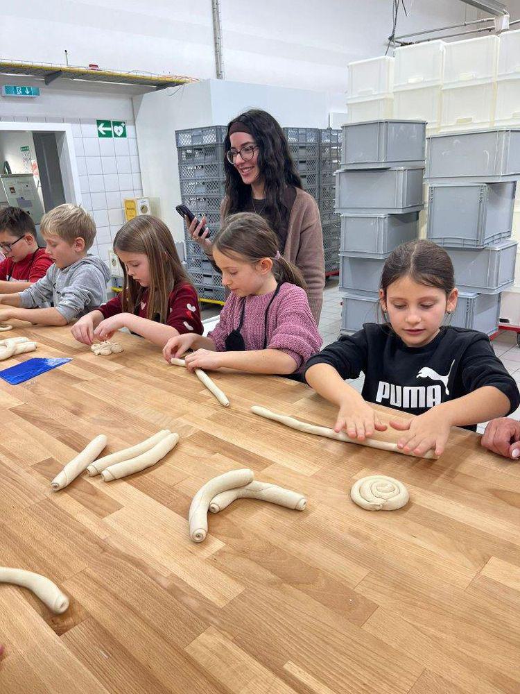 Mehrere Kinder sitzen an einem Holztisch und rollen Teig zu langen Rollen. Im Hintergrund stehen gestapelte graue Kunststoffkisten.