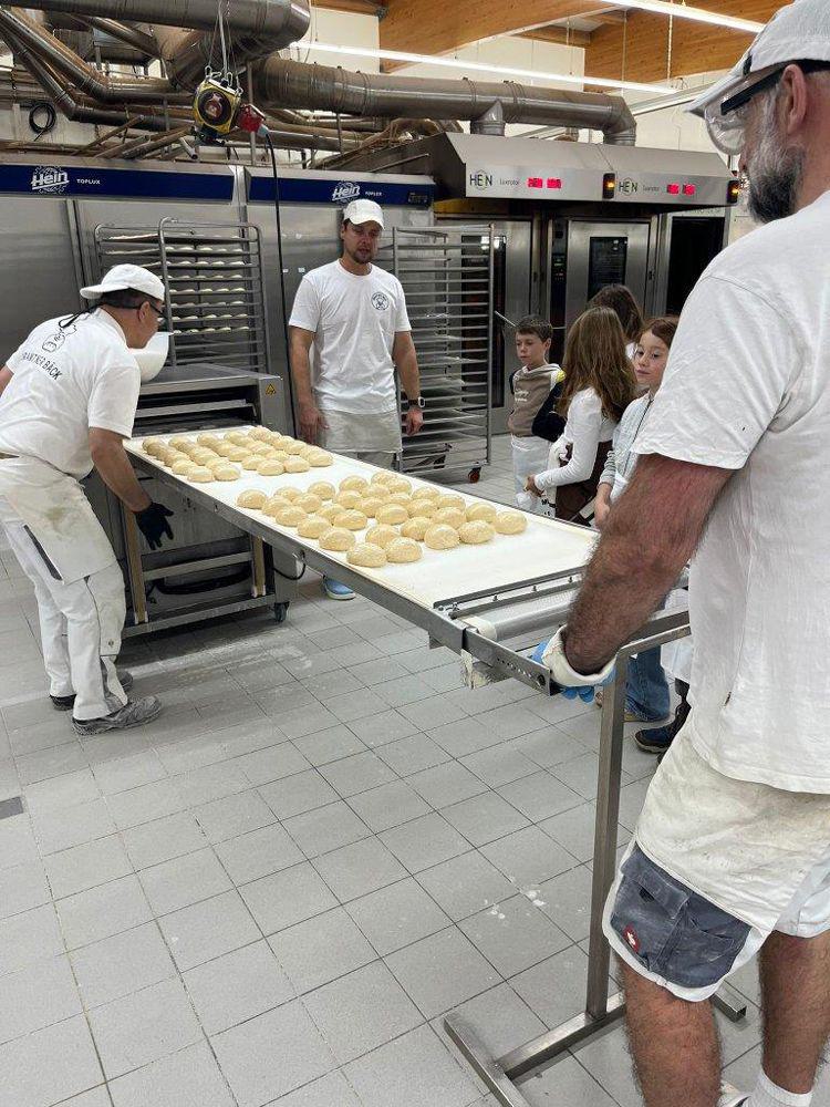 Drei Bäcker in weißer Arbeitskleidung tragen ein großes Tablett mit mehreren runden Teiglingen in einer Bäckerei mit Backöfen im Hintergrund.