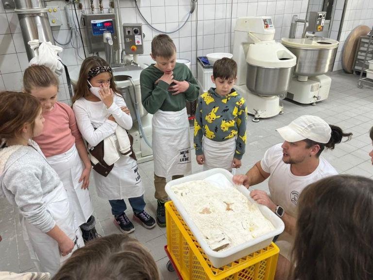 Mehrere Kinder mit Schürzen stehen in einer Bäckerei um eine mit Teig gefüllte Wanne, ein Mann in weißer Kleidung erklärt etwas.