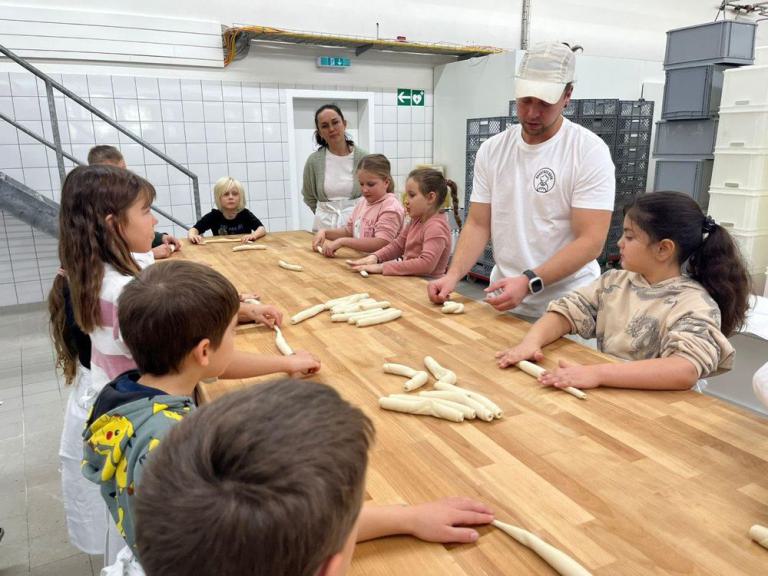 Mehrere Kinder sitzen an einem langen Holztisch, ein Mann mit weißer Kochmütze und weißem T-Shirt zeigt ihnen längliche Teigstücke.