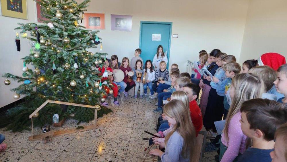 Kinder vor einem geschmückten Weihnachtsbaum, einige spielen Glockenspiele, andere halten Liederbücher.