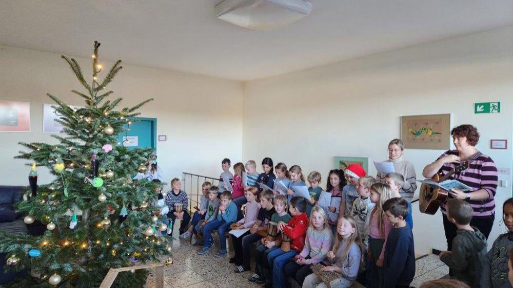 Weihnachtsbaum mit Kugeln und Lichterkette, davor sitzende Kinder und zwei Erwachsene, einer spielt Gitarre.