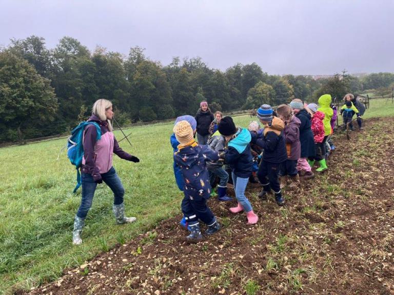 Gruppe von Kindern mit bunter Kleidung zieht gemeinsam an einem Seil auf einem Feld, begleitet von zwei Erwachsenen.