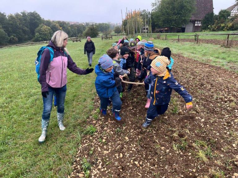 Gruppe von Kindern in bunter Outdoor-Kleidung zieht gemeinsam an einem Seil auf einem Feld, zwei Erwachsene begleiten sie.