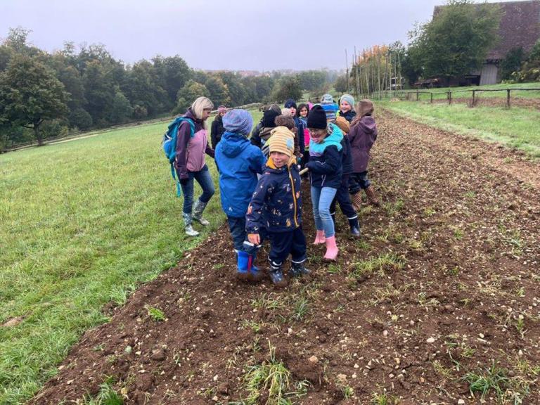 Gruppe von Kindern mit bunter Kleidung und Gummistiefeln steht auf einem Acker neben einer Wiese, im Hintergrund Bäume und ein Haus.