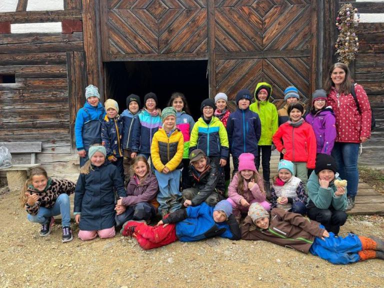 Gruppe von Kindern in bunter Outdoor-Kleidung vor einem großen Holztor mit diagonalem Muster, einige sitzen, andere stehen.