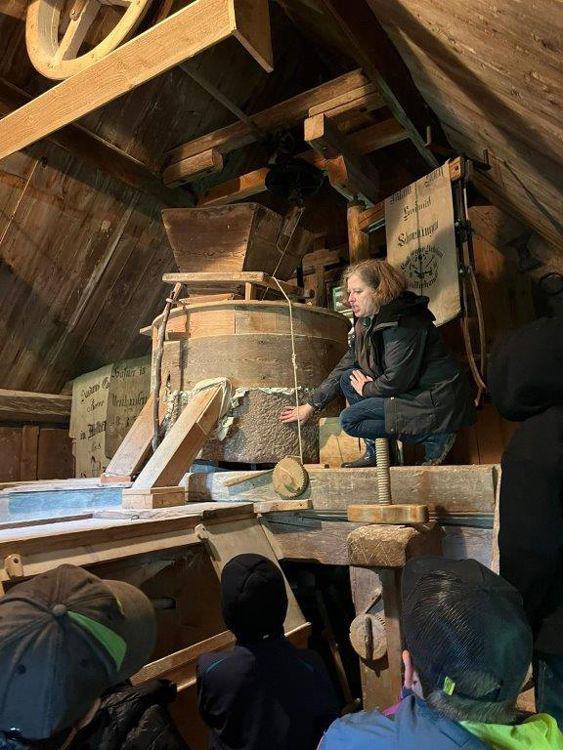 Eine Frau kniet bei einer historischen Mühle und erklärt Kindern die Funktion