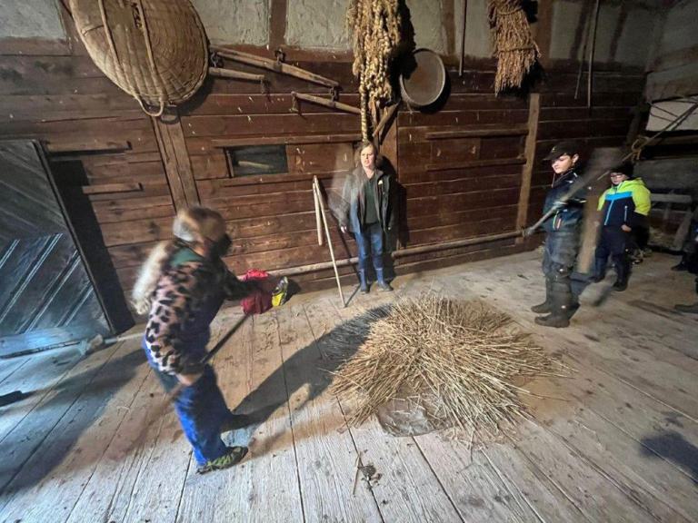 Zwei Kinder dreschen Getreide auf einem Holzboden, eine Frau und ein weiteres Kind schauen zu