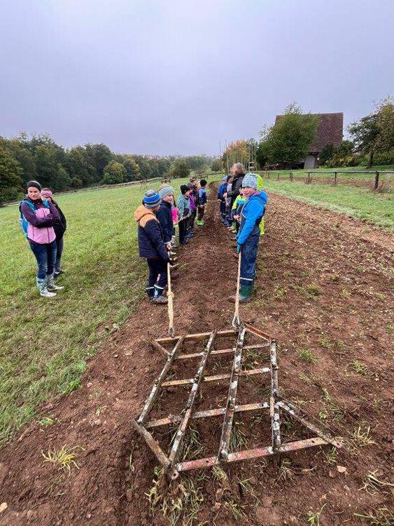 Kinder stehen in zwei Reihen auf einem frisch gepflügten Feld, vorne liegt ein hölzernes Pfluggerät mit zwei langen Griffen.