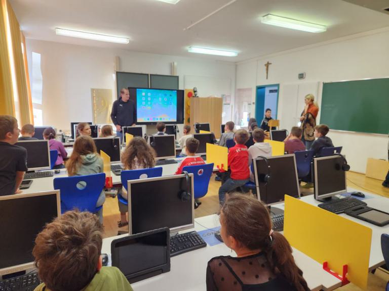 Schulklasse sitzt an Computertischen, vorne steht ein Lehrer neben einem großen Bildschirm mit Windows-Desktop, rechts Lehrerin vor Tafel.