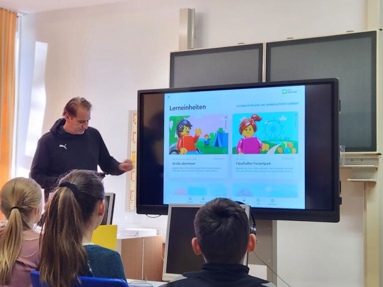 Lehrer zeigt auf Bildschirm mit zwei Lernmodulen: 'Große Abenteuer' und 'Fabelhafter Freizeitpark', mehrere Schüler sitzen davor.