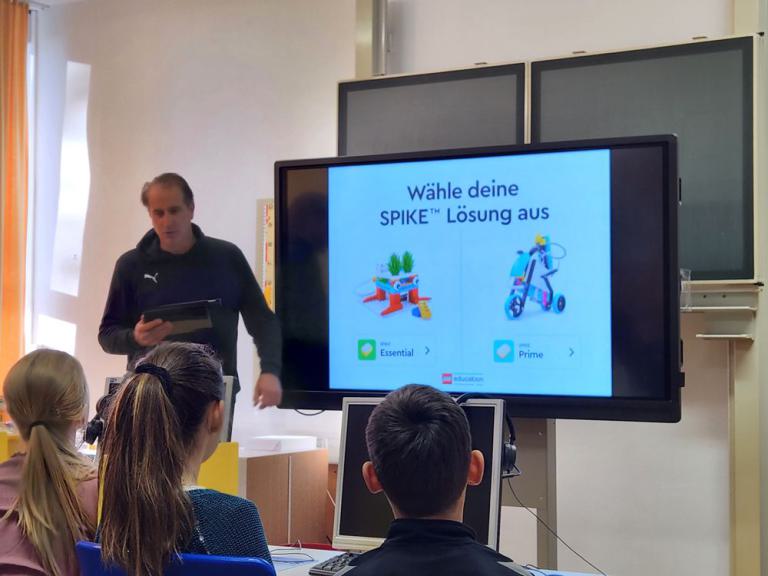 Mann steht vor Bildschirm mit Präsentation zu SPIKE-Lösungen, mehrere Personen sitzen und schauen zu.