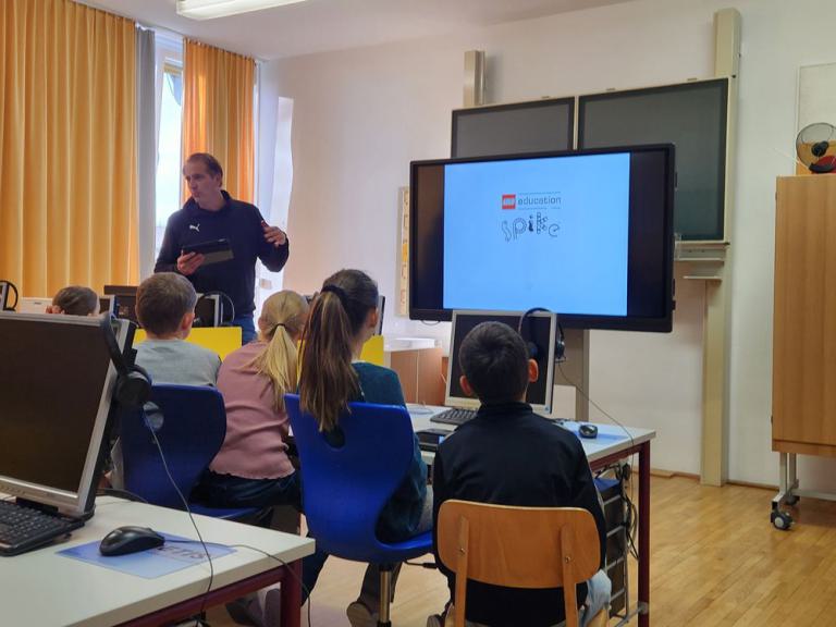Lehrer steht vor Klasse mit mehreren Kindern, die auf Stühlen vor einem Bildschirm sitzen, auf dem 'education spike' zu sehen ist.