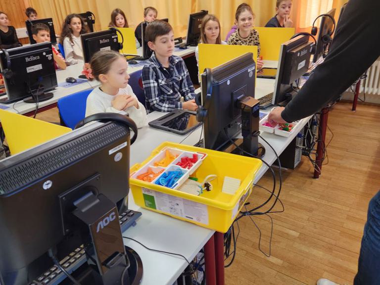 Mehrere Kinder sitzen an Computertischen mit Monitoren und gelben Kisten voller bunter Bauteile, eine Person steht rechts und hält einen Gegenstand.