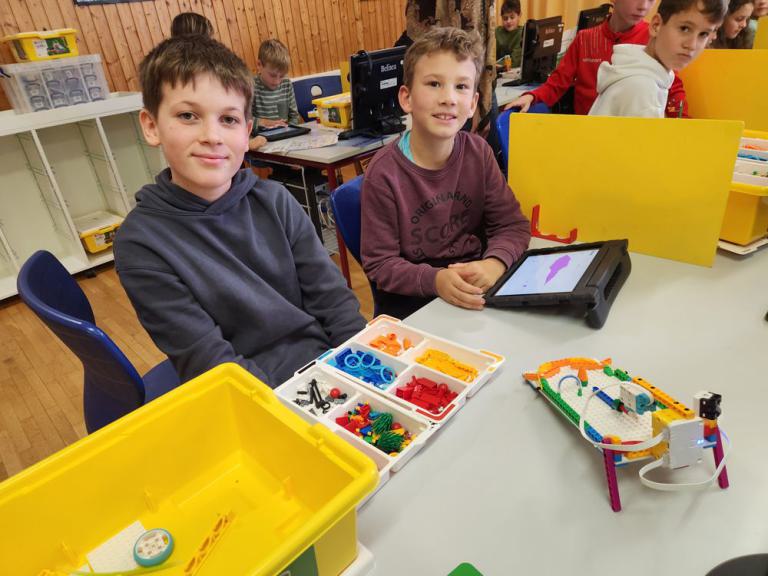 Zwei Jungen sitzen an einem Tisch mit einem Tablet, einem Sortierkasten mit bunten Bauteilen und einem kleinen Roboter aus Bausteinen.