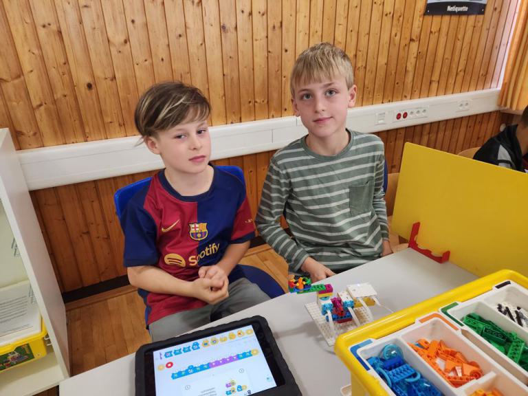 Zwei Jungen sitzen an einem Tisch mit einem Tablet und einem kleinen Roboter aus bunten Bausteinen.