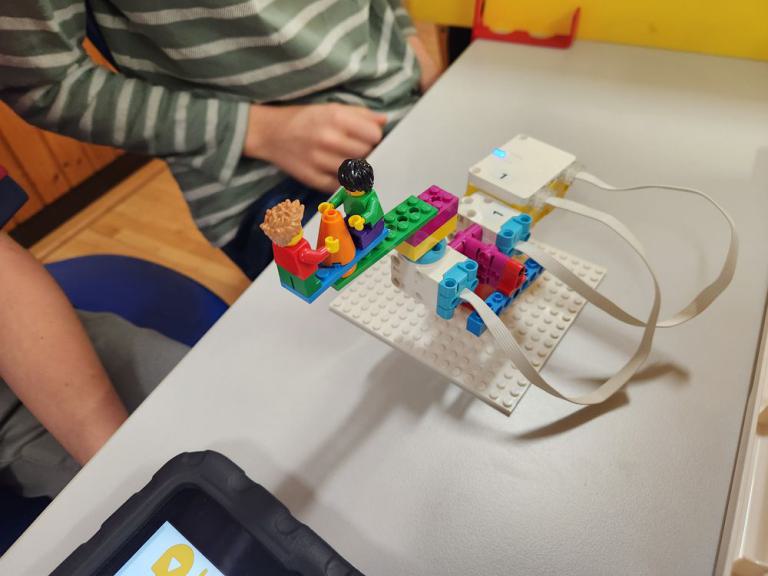 Bunte Lego-Figuren auf einer kleinen Lego-Plattform, verbunden mit weißen Kabeln an ein weißes Gerät auf einem Tisch.