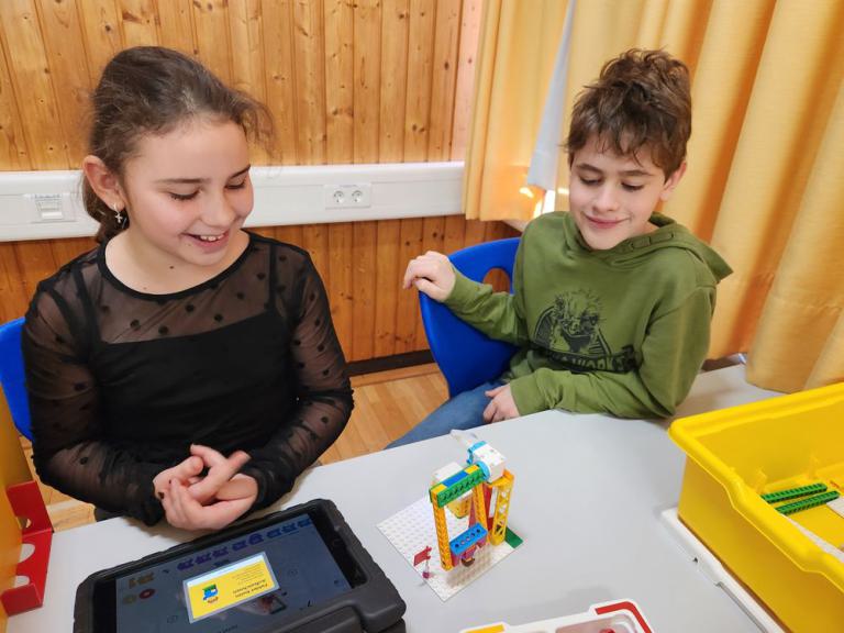 Zwei Kinder sitzen an einem Tisch mit einem Tablet und einem bunten Lego-Roboter-Modell auf einer weißen Bauplatte.