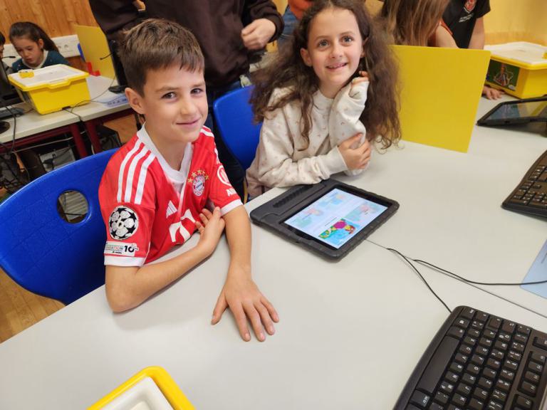 Zwei Kinder sitzen an einem Tisch, eines trägt ein rotes Trikot mit FC-Bayern-Logo, auf dem Tisch liegt ein Tablet mit bunter Anzeige.