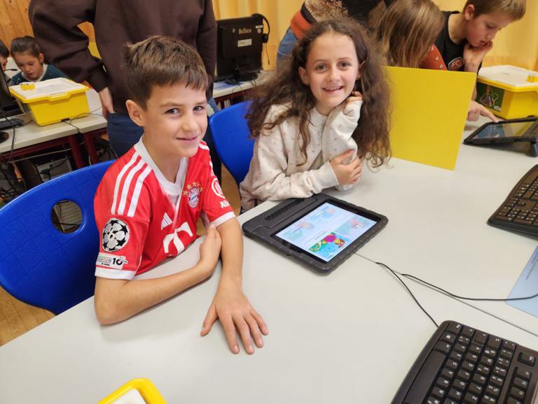 Zwei Kinder sitzen an einem Tisch, eines trägt ein rotes Fußballtrikot, vor ihnen liegt ein Tablet mit bunter Anzeige, im Hintergrund weitere Kinder und gelbe Boxen.