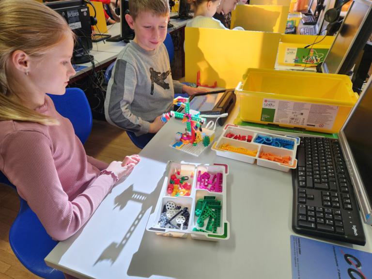 Zwei Kinder sitzen an einem Tisch mit bunten Bauteilen und einem kleinen Roboter aus Legosteinen, daneben Tastatur und Bildschirm.