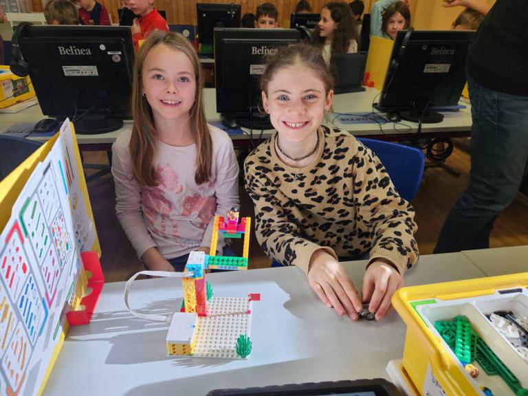 Zwei Kinder sitzen an einem Tisch mit einem kleinen Roboter aus bunten Bausteinen und einem gelben Kasten mit weiteren Bausteinen.