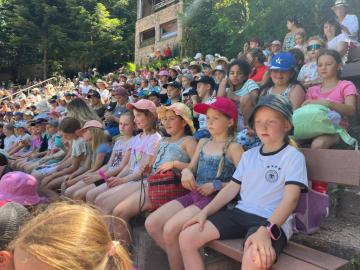 Viele Kinder mit bunten Basecaps sitzen an einem sonnigen Tag aufmerksam auf den Bänken eines voll besetzten Outdoor-Amphitheaters.