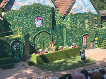 Eine bunte, fantasievolle Szenerie mit einem grünen, niedrig gehaltenen Podest, umgeben von gemalten Wänden und einem kunstvollen Eingang.