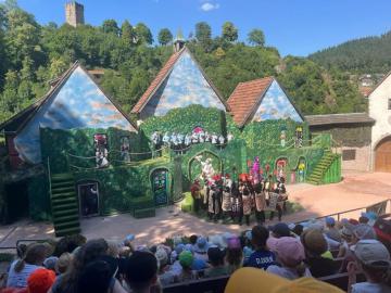 Eine bunte, theatralische Kulisse, die ein Märchenhaus oder einen Dorfkomplex darstellt, umgeben von grünen Hügeln und beobachtet von einer Gruppe von