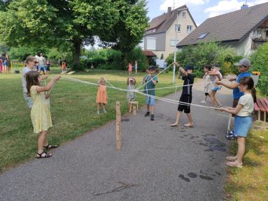 Kinder und Erwachsene spielen ein Seilspiel auf einem asphaltierten Weg in einer Grünanlage vor Wohnhäusern.