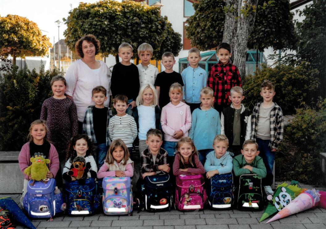 Ein Gruppenbild mit SchülerInnen und Lehrerin in drei Reihen, die vorderste Reihe hat Ranzen von sich stehen. Zei Schultüten liegen daneben.