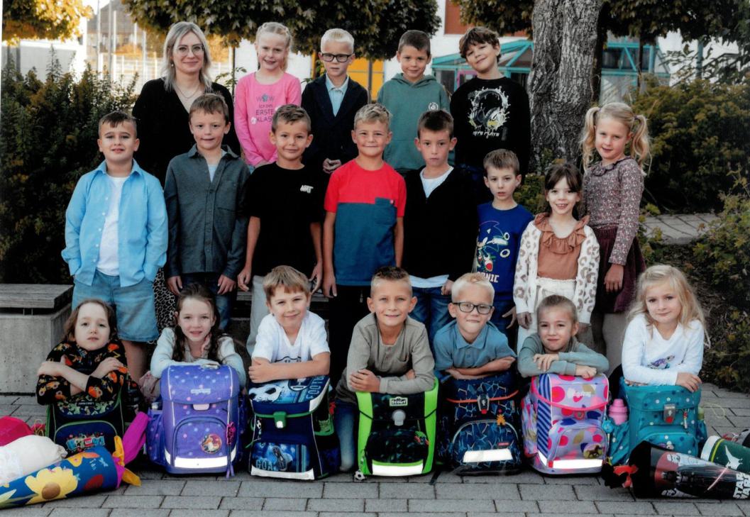 Eine Gruppenbild von SchülerInnen und Lehrerin in drei Reihen, die vordertste Reihe hat Schulranzen vor sich stehen